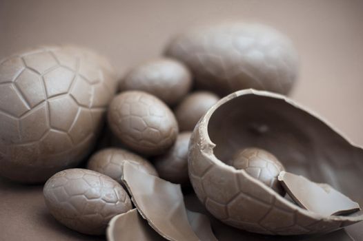 Brown background with a broken chocolate Easter egg in the foreground and an assortment of whole eggs behind with shallow dof