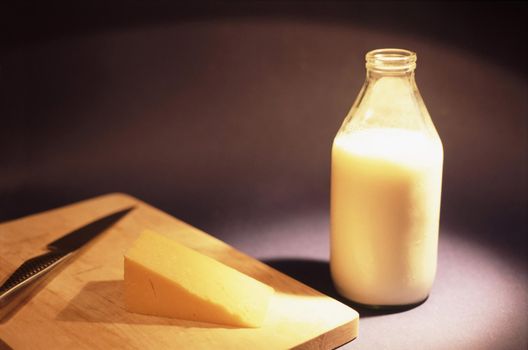 Glass bottle of fresh cows milk standing alongside a wedge of cheddar cheese on a cheeseboard against a dark background with copyspace