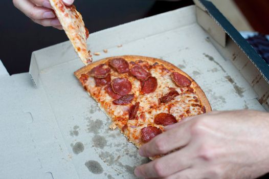 Hands of a man eating a slice of a takeaway pepperoni pizza still in its traditional cardboard box