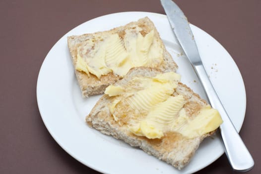 Close up Tasty Toasted Bread with Butter on Top Served on White Round Plate with Knife. Isolated on Brown Background.