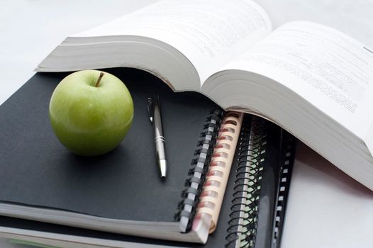 Conceptual background for education and college studies with a large open textbook alongside spiral bound notebooks and a pencil topped with a fresh healthy green apple