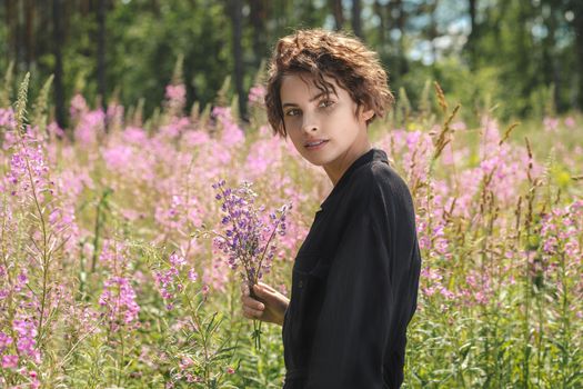 Young woman with a bouquet of lupines in her hand against the background of blooming Ivan-tea on a bright sunny summer day
