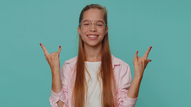 Overjoyed delighted pretty girl showing rock n roll gesture by hands, cool sign, shouting yeah with crazy expression, dancing, emotionally rejoicing in success. Young teen child kid on blue background