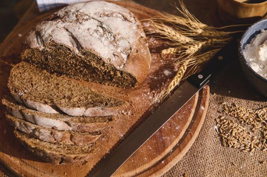Still life with sliced loaf of a homemade whole grain sourdough rye bread, a kitchen knife on a wooden cutting board, and wheat ears near a bowl with white flour and scattered cereal grains on burlap