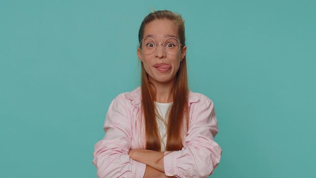 Cheerful funny bully teenager girl in shirt. Child girl showing tongue making faces at camera, fooling around, joking, aping with silly face, teasing isolated alone on blue studio background indoors
