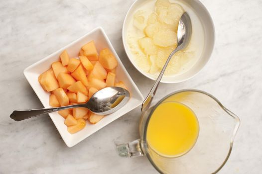 Variety of breakfast fruits with orange juice in a jug, and fresh diced melon and pears in individual dishes, overhead view