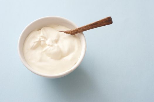 Small white round bowlk of plain yogurt with wooden spoon against a light colored background