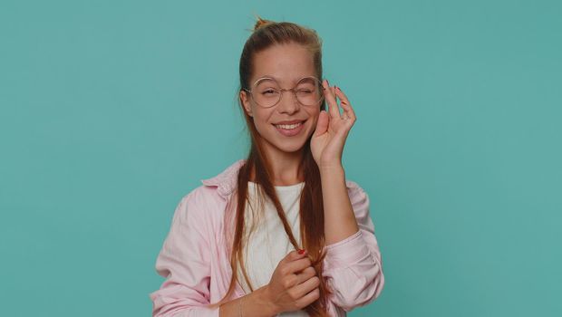 Smiling playful happy girl blinking eye, looking at camera with toothy smile, winking and flirting, expressing optimism. Young stylish female child kid isolated alone on blue studio background indoors
