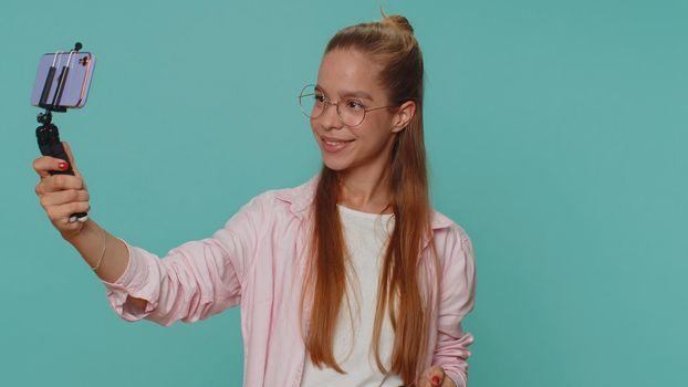 Pretty teenager girl traveler blogger taking selfie on mobile phone selfie stick, communicating video call online with subscribers. Young child kid isolated alone on blue studio background indoors