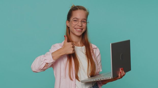Girl working online on laptop, finishing work, showing thumb up to camera, positive feedback, like, lottery win, approve, good offer advertisement. Young teen child kid on blue studio wall background