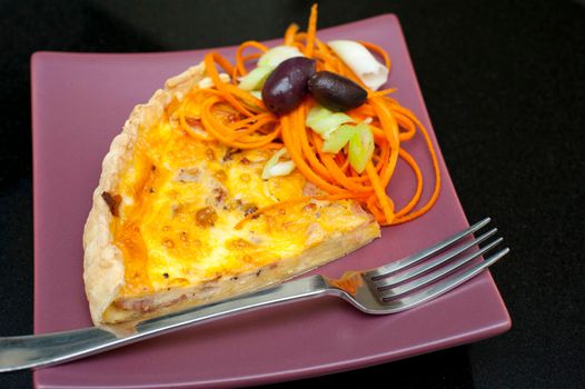 Slice of savoury quiche baked with an egg custard base and served with a side portion of spiced spaghetti and olives