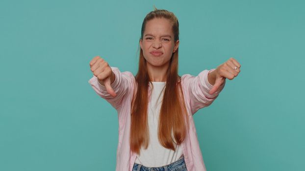 Upset lovely pretty teenager girl showing thumbs down sign gesture, expressing discontent, disapproval, dissatisfied, dislike. Young stylish female child kid isolated alone on blue studio background