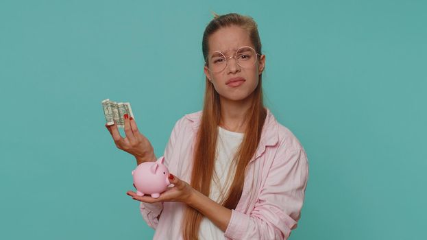 Poor pretty teenager girl insufficient amount of money, holding piggybank and one dollar banknote. Financial crisis. Bankruptcy. Poverty and destitution. Young child kid on blue background indoors