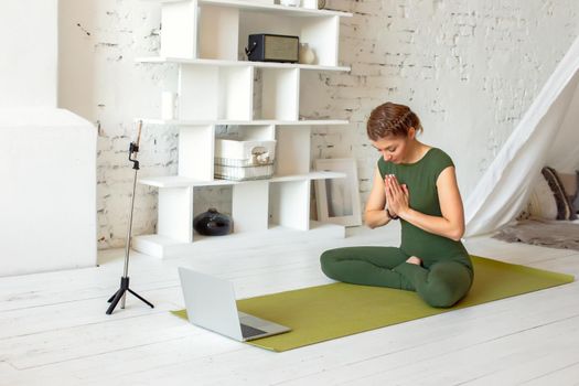 A slender beautiful woman in a green sports jumpsuit, in a bright room, is broadcasting online, performs a yoga exercise, there is a laptop and a smartphone on a tripod nearby