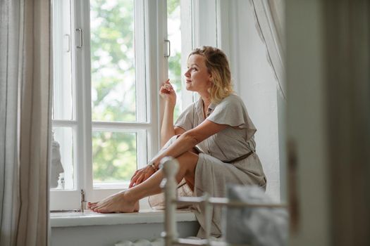 A middle-aged woman in a cream dress sits mysteriously and looks out the window on the windowsill. Green trees outside