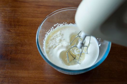 Whipping cream in a glass mixing bowl with a whisk attachment on an electric kitchen mixer, high angle close up view