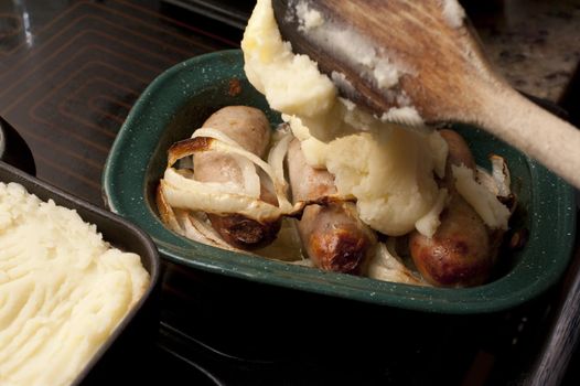 Preparing sausage pie in the kitchen with a close up view of the mashed potato topping being applied with a wooden spoon