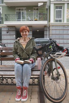 Vertical shot of a happy young woman laughing, reading a book in city center, resting after cycling