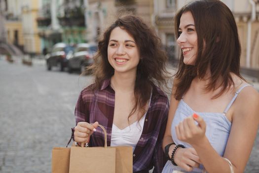 Happy young female friends laughing, walking city streets, carrying shopping bags. Vacation, sighseeing, traveling concept