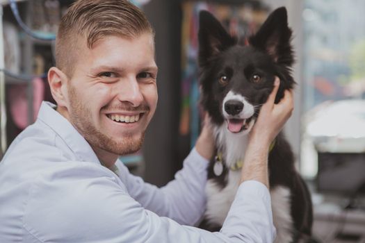 Handsome happy male veterinarian laughing to the camera, petting adorable dog at his clinic. Cute black puppy being examined by professional vet doctor. Happiness, animals, love concept