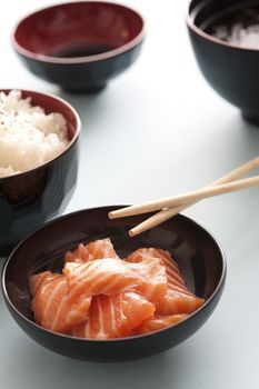 Fresh salmon dinner with chopsticks in black bowl besides serving of white rice and soy dipping sauce