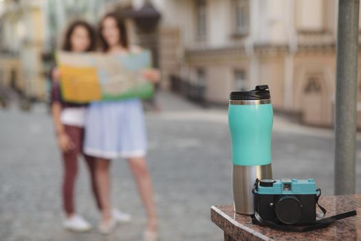 Selective focus on a vintage camera and thermo cup, female friends using map on the background. Women sightseeing while travelling together