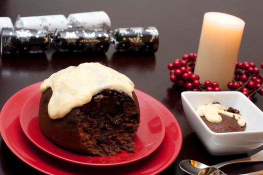 Serving of traditional steamed fruity Christmas pudding and brandy custard on a festive table with a glowing seasonal candle and wreath of colourful red berries