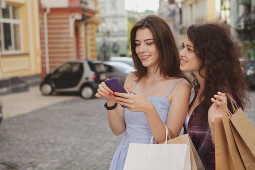 Two lovely young women using smart phone, walking in the city after shopping together, copy space