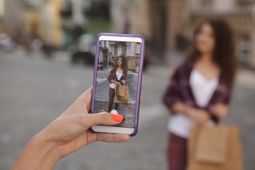 Cropped close up of a woman using smart phone, taking photos of her female friend in the city. Female friends wandering on city streets, taking photos with mobile phone