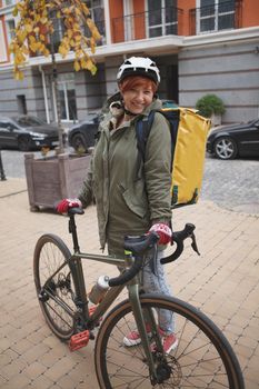 Vertical full length shot of a cheerful young woman working as delivery courier in the city, using her bicycle
