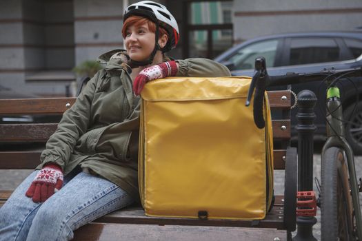 Young happy female courier wearing bicycle helmet, resting after cycling city streets with her delivery backpack