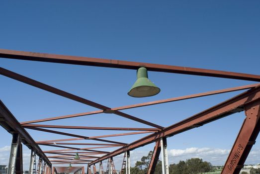 Overhead rusty girders of a bridge structure with an electric light with shade in an open framework against a blue sky