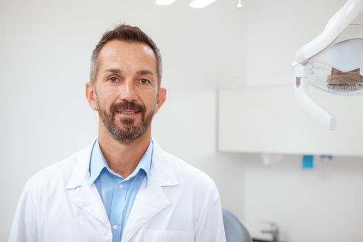 Cheerful mature male dentist smiling to the camera at his office, copy space