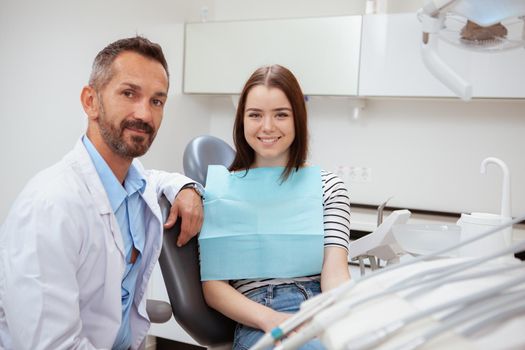 Attractive young woman sitting in a dental chair next to her dentist, copy space. Mature dentist and his female patient smiling to the camera after dental checkup