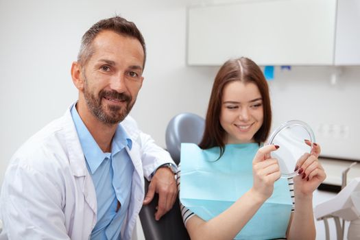 Handsome mature male doctor smiling to the camera, his female patient smiling looking in the mirror. Young woman checking her teeth in the mirror after professional dental treatment