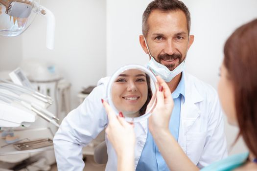 Cheerful mature male dentist smiling to the camera, his female patient checking her teeth in the mirror after dental treatment