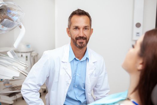 Happy handsome mature male dentist smiling to the camera while working with a patient at his clinic