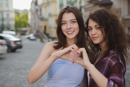 Happy young women smiling to the camera, showing a heart with their hands, copy space. Lovely female friend holding their hands in a heart shape. Love, friendship, happiness; concept