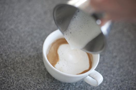 Adding steamed frothy milk to a cup of freshly brewed cappuccino coffee, closeup of a hand pouring the milk from a metal jug, focus to the coffee