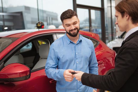 Charming bearded man receiving car keys from the salesman after buying a new automobile, copy space. Deal, contract, rental concept