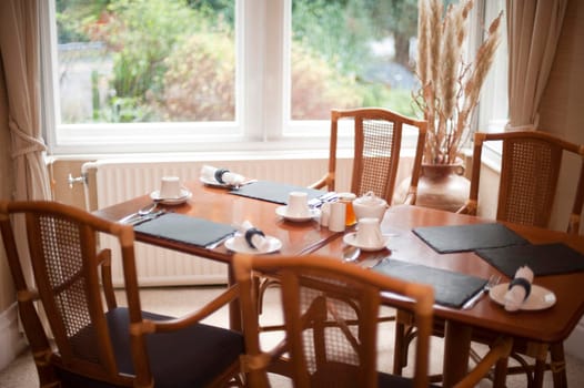 Hardwood dining table set for three people with stylish wicker back chairs in front of a bright window