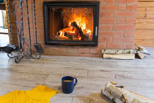 Fireplace with a blazing fire in the interior of a wooden house. Firewood and a fireplace set are located near the hearth with a flame..