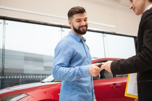 Low angle shot of a handsome bearded man receiving keys to his new car at the automobile dealership, copy space. Car buying, ownership concept