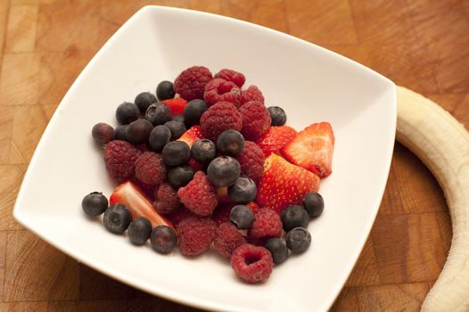 Bowl of fresh autumn fruit with assorted berries including strawberries, blueberries and raspberries with a peeled whole banana alongside the dish