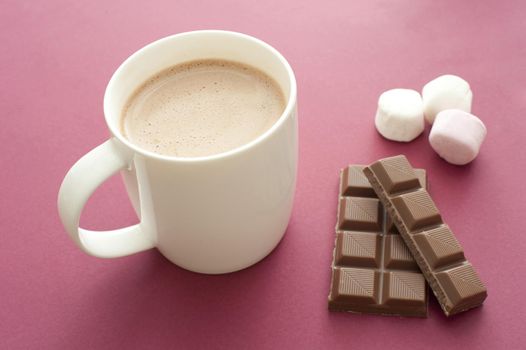 Delicious hot chocolate drink in a mug with ingredients alongside including a bar of chocolate candy and marshmallows, on a red background, high angle view