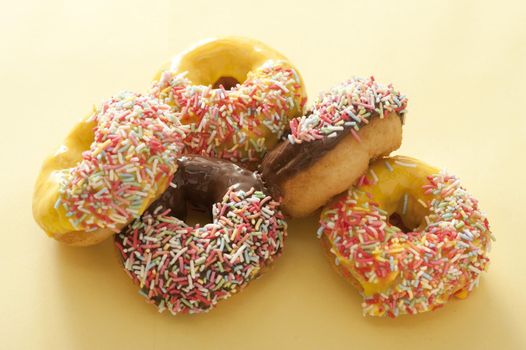 Pile of assorted fresh ring donuts or doughnuts glazed with chocolate, lemon or orange icing decorated with colorful sprinkles on a pale yellow background