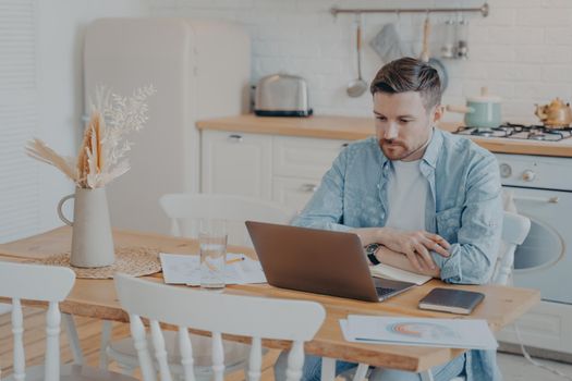 Serious and focused young freelancer working on laptop while sitting by kitchen table with hands resting on it, graphs and statistics scattered around, open note book next to him