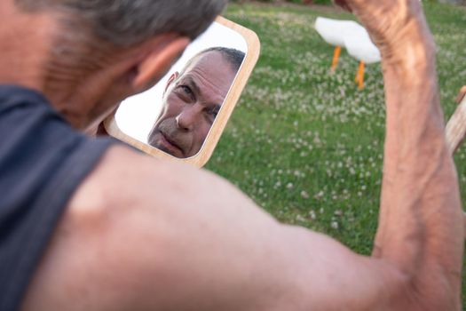 a gray-haired middle-aged man shaves his hair with a clipper in a garden on a green lawn and looks into a table mirror. High quality photo