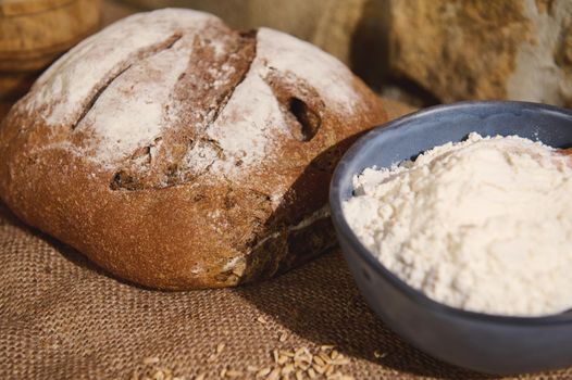 Selective focus on fresh baked wholesome homemade traditional wheaten sourdough bread, and blurred blue ceramic bowl with white flour on burlap and scattered grains of wheat on the blurred foreground