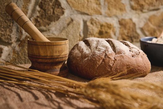 Still life. Food composition with a loaf of traditional whole grain homemade wholesome sourdough wheat bread, on a linen tablecloth next to a wooden mortar and wheat spikelets in blurred foreground